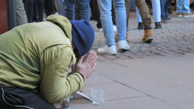 A poor homeless man begs for alms on his knees on the street in winter with his hands folded in a begging gesture. Crowds of people pass by slow motion