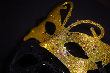 A close-up photo of two carnival masks, one gold and one black, on a black background. The gold...