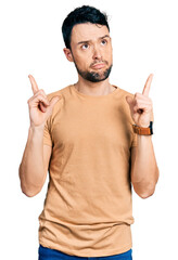 Hispanic man with beard wearing casual t shirt pointing up looking sad and upset, indicating direction with fingers, unhappy and depressed.