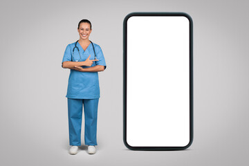 Cheerful european woman nurse, surgeon in blue uniform point at big phone with empty screen, isolated on gray background