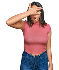 Fototapeta premium Young hispanic girl wearing casual t shirt peeking in shock covering face and eyes with hand, looking through fingers with embarrassed expression.