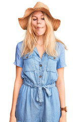 Fototapeta premium Young beautiful blonde woman wearing summer hat and dress making fish face with lips, crazy and comical gesture. funny expression.