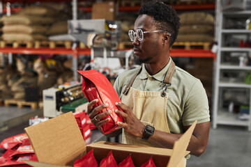 Waist up portrait of Black young man holding coffee bag doing quality control in packaging...