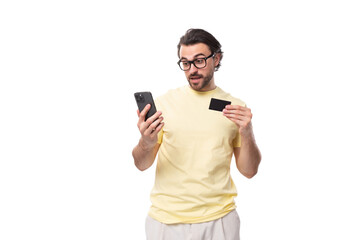 casual caucasian guy with a beard in glasses dressed in a light t-shirt uses a smartphone and a plastic card