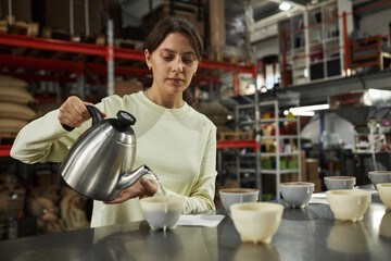 Waist up portrait of young woman pouring hot water into small bowls during cupping and quality control inspection at coffee factory, copy space