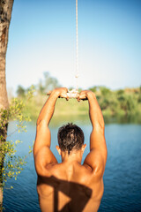 Unrecognizable muscular young guy seen from behind thinking about jumping into water from a rope/zip line. Caucasian boy doing water sport.