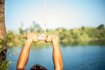 Detail of hands of young Caucasian man grabbing a stick and a rope tied to a tree to jump into the water in a lake. Sunny summer day.