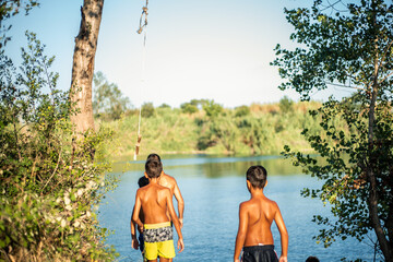 A group of unrecognizable young boys seen from behind thinking about jumping with a rope into the water of a lake in summer.