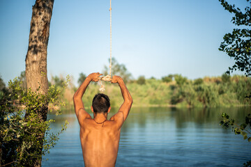 Unrecognizable muscular young guy seen from behind thinking about jumping into water from a rope/zip line. Caucasian boy doing water sport.