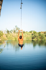 Unrecognizable muscular young guy seen from behind jumping into the water from a rope/zip line while doing a flip. Caucasian boy doing water sport.