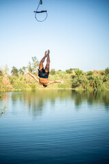 Unrecognizable muscular young guy seen from behind jumping into the water from a rope/zip line while doing a flip. Caucasian boy doing water sport.