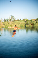Unrecognizable muscular young guy seen from behind jumping into the water from a rope/zip line while doing a flip. Caucasian boy doing water sport.