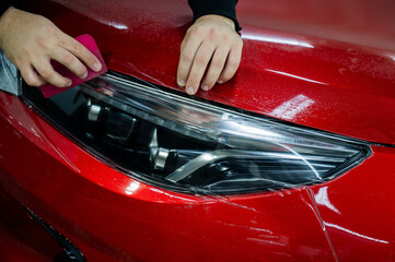 The master applies vinyl film to the headlight of a red car. Closeup view on worker detailer hand...