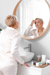 Fototapeta premium Mature woman applying facial cream near mirror in bathroom