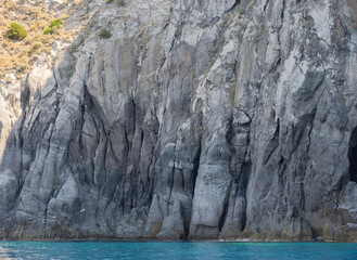 Weathered seaside rock face texture with parts of green and blue water. Aged volcanic stone wall surface background pattern with cracks and scratches. Ischia Island, Italy.