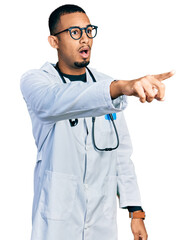 Young african american man wearing doctor uniform and stethoscope pointing with finger surprised...