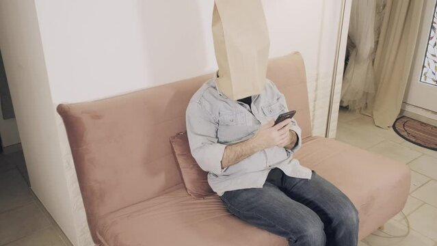 A man wearing a breadbag face, sitting on the sofa at home, no expression on his paper mask; checking his smartphone, bored, throwing it away.
