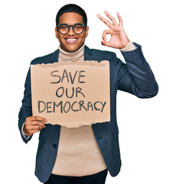 Young handsome hispanic man holding save our democracy protest banner doing ok sign with fingers, smiling friendly gesturing excellent symbol