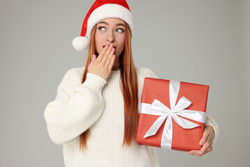Emotional young woman in Santa hat with Christmas gift on light grey background
