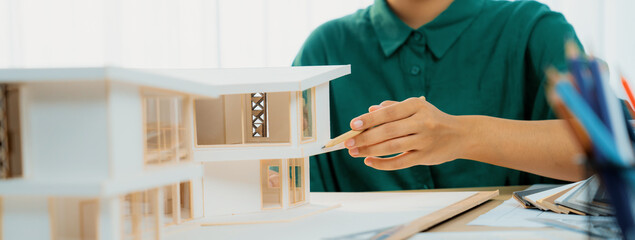 A cropped imaged of female designer measures a house model with a pencil, focusing intently on her...