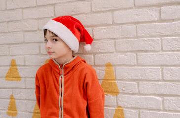 a little boy in orange pajamas and a red Santa Claus hat looks suspiciously at the camera against...