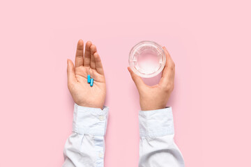 Female hands with blue pills and glass of water on pink background