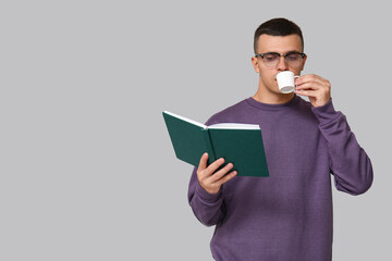 Handsome young man reading book and drinking coffee on grey background