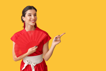 Beautiful young happy woman with fan pointing at something on yellow background. Chinese New Year...