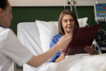 Young physician sits with senior patient in hospital room, discussing and obtaining surgical...