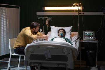Young girl seriously ill lying on a bed dressed in an oxygen mask man sitting on the side girl after an accident unconscious in a coma. - obrazy, fototapety, plakaty
