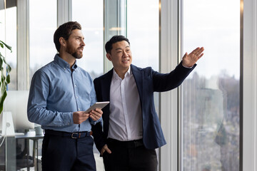 Two business colleagues are standing by the window in the office, looking at the view from the...