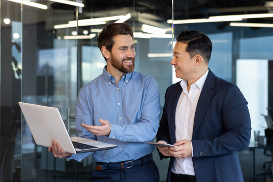 Two businessmen men of different races are standing in the office, looking at a laptop, discussing work topics, smiling at each other.
