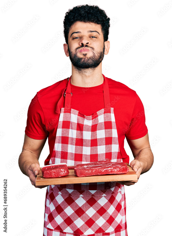 Poster young arab man with beard holding board with raw meat puffing cheeks with funny face. mouth inflated