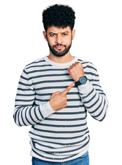 Young arab man with beard wearing casual striped sweater in hurry pointing to watch time,...
