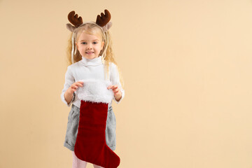 Cute little girl in reindeer horns with Christmas sock on beige background