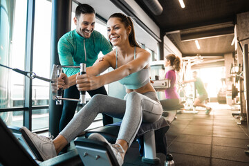 Fototapeta na wymiar Happy female athlete doing rowing workout with personal trainer in the gym. Cheerful fitness coach showing seated cable rows exercise to his client.