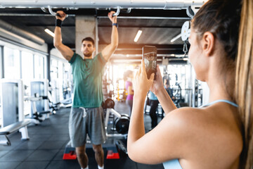Young sportswoman taking a picture of athlete doing exercise in the gym. Rear view of a young woman...