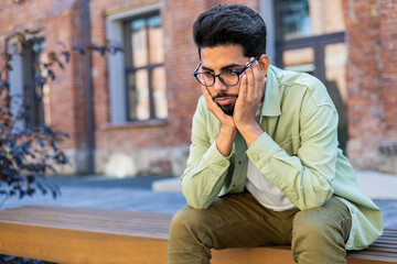 An upset Indian man in glasses sits on a street bench, holds his head in his hands, sad news, problems at work.