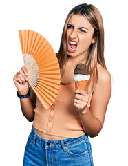 Hispanic young woman holding hand fan eating ice cream angry and mad screaming frustrated and...