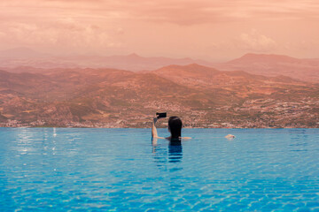 Woman swimming in the pool water and taking a pucture on the phone of mountaun with Greek village. Swimming pool on the roof. Summer recreation