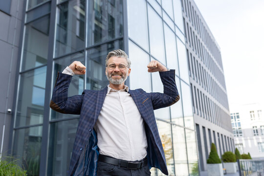Successful senior businessman in suit standing in front of office building, raising hands up, flexing muscles, smiling and looking at camera.