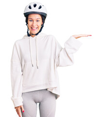 Beautiful brunette young woman wearing bike helmet and sporty clothes smiling cheerful presenting and pointing with palm of hand looking at the camera.