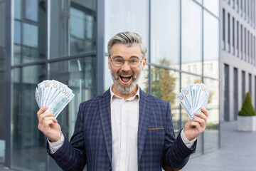 Businessman is delighted with a huge win, holds stacks of dollars and shouts with joy, looks at the...