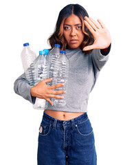 Young beautiful mixed race woman holding recycling plastic bottles with open hand doing stop sign with serious and confident expression, defense gesture