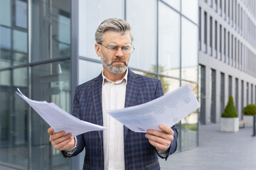 Angry senior boss standing on the street in front of the office, holding work documents, problems at work, bad news.