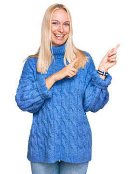 Young blonde girl wearing wool winter sweater smiling and looking at the camera pointing with two hands and fingers to the side.