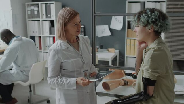 Supervising physician holding digital tablet checking on her young patient with amputation while her colleague working on computer in background