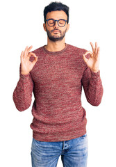 Young handsome hispanic man wearing winter sweater and glasses relax and smiling with eyes closed doing meditation gesture with fingers. yoga concept.