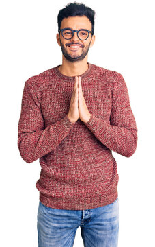 Young handsome hispanic man wearing winter sweater and glasses praying with hands together asking for forgiveness smiling confident.