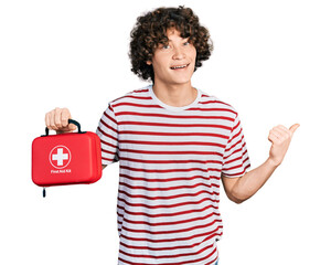 Caucasian teenager holding first aid kit pointing thumb up to the side smiling happy with open mouth